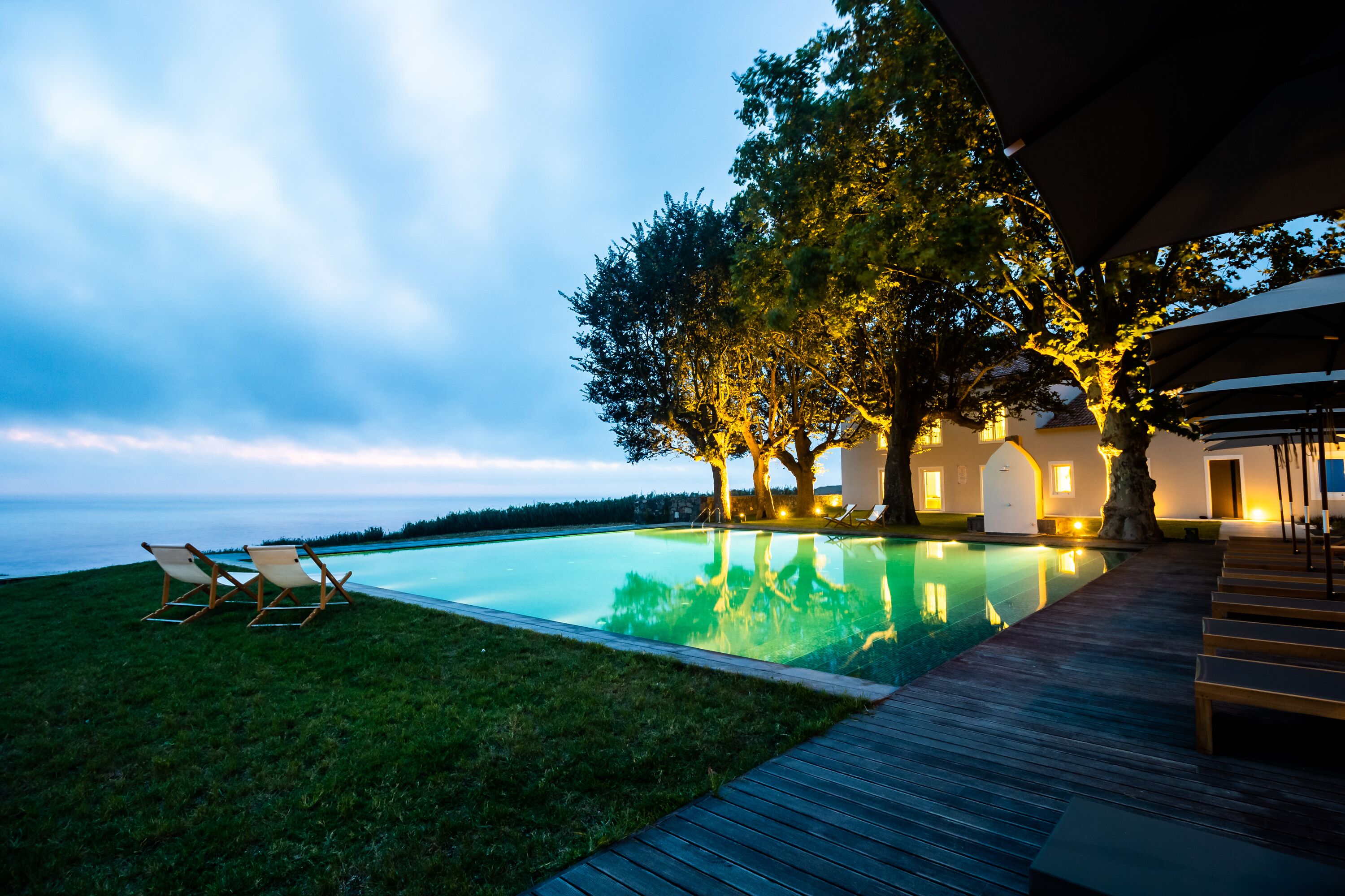 Une piscine entourée d’arbres, avec des chaises longues et une terrasse en bois, et un bâtiment dont l’éclairage est chaleureux.