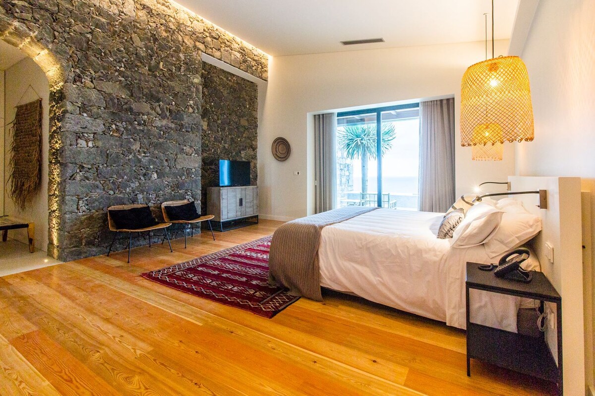 A bedroom with a stone wall, a bed, a TV, a desk, and a view of the outdoors.