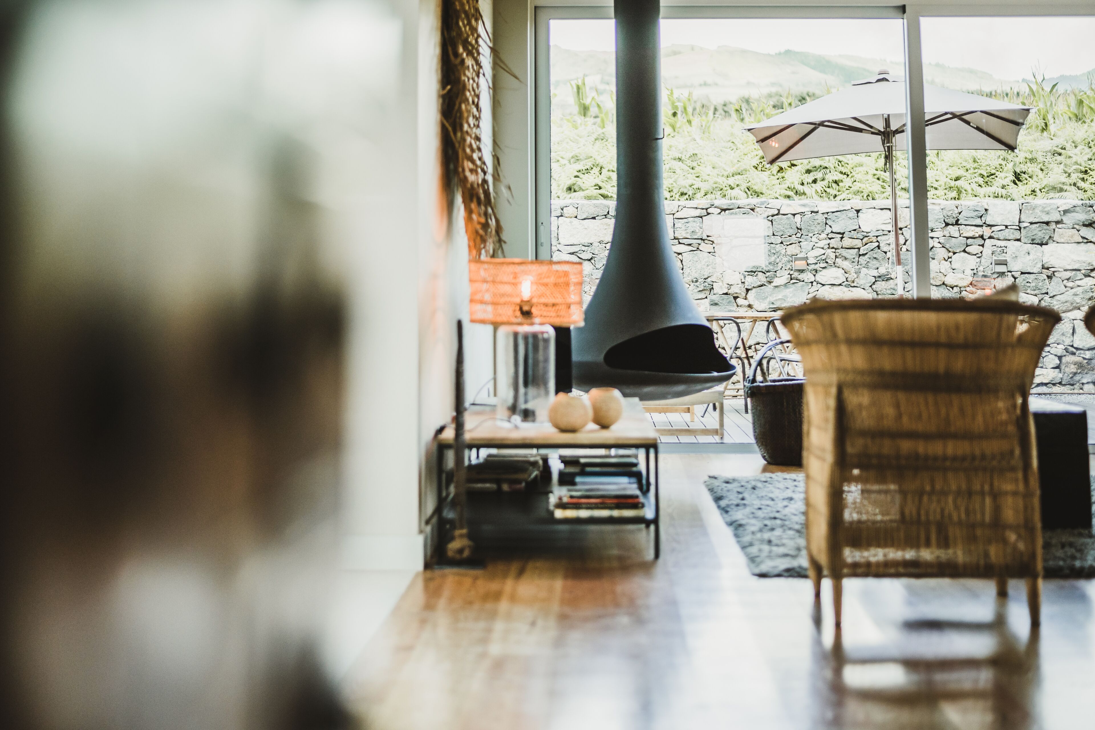 A cozy living room with a fireplace, wicker furniture, and a view of a stone wall and greenery outside.