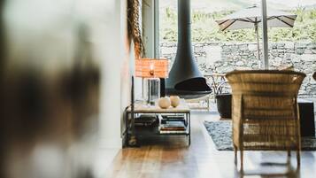 A cozy living room with a fireplace, wicker furniture, and a view of a stone wall and greenery outside.
