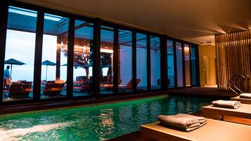 An indoor swimming pool with clear glass walls, lounge chairs, and a wooden deck.