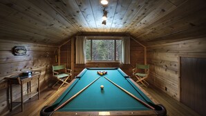 Um quarto de madeira com mesa de bilhar, duas cadeiras, uma janela e um relógio na parede.