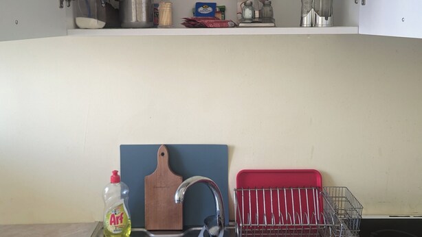 Een keuken met een spoelbak, een afdruiprek en bovenkasten met keukengerei en etenswaren.