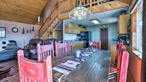 A spacious dining area with a long wooden table, red chairs, and a chandelier. A kitchen is visible in the background with wooden cabinets and a stove. - Ruidoso Home w/ Hot Tub, Mtn Views & Game Room! (Ruidoso)