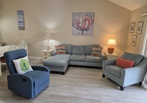 A living room with a sectional sofa, a blue armchair, a side table with a lamp, and a painting on the wall.