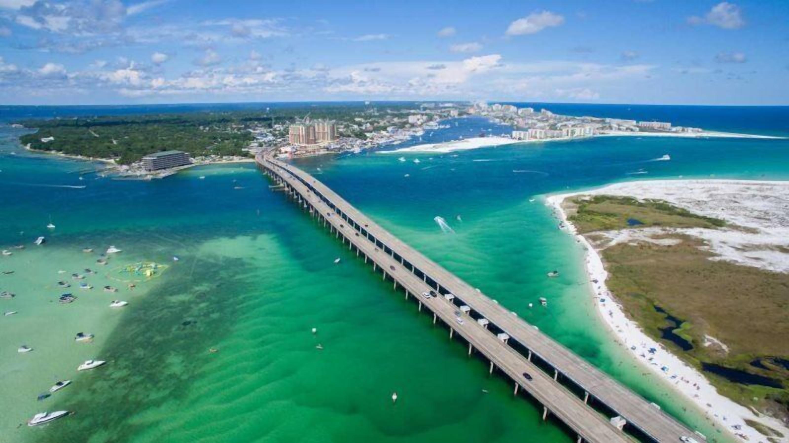 A coastal bridge with clear turquoise waters, a beach, and a cityscape in the background.