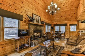 A wooden cabin interior with a stone fireplace, a television, a dining area, and a cozy seating arrangement.