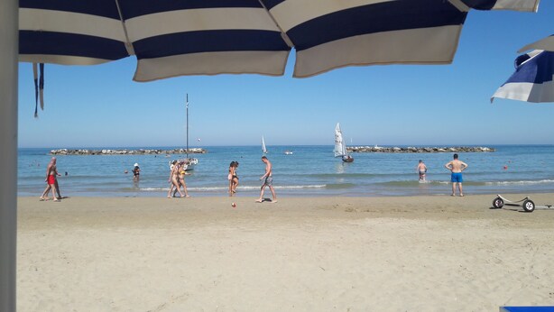 Escena de playa con gente caminando y un velero en el agua.