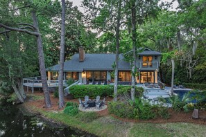Une maison à deux étages avec une terrasse, entourée d’arbres et d’un petit étang.
