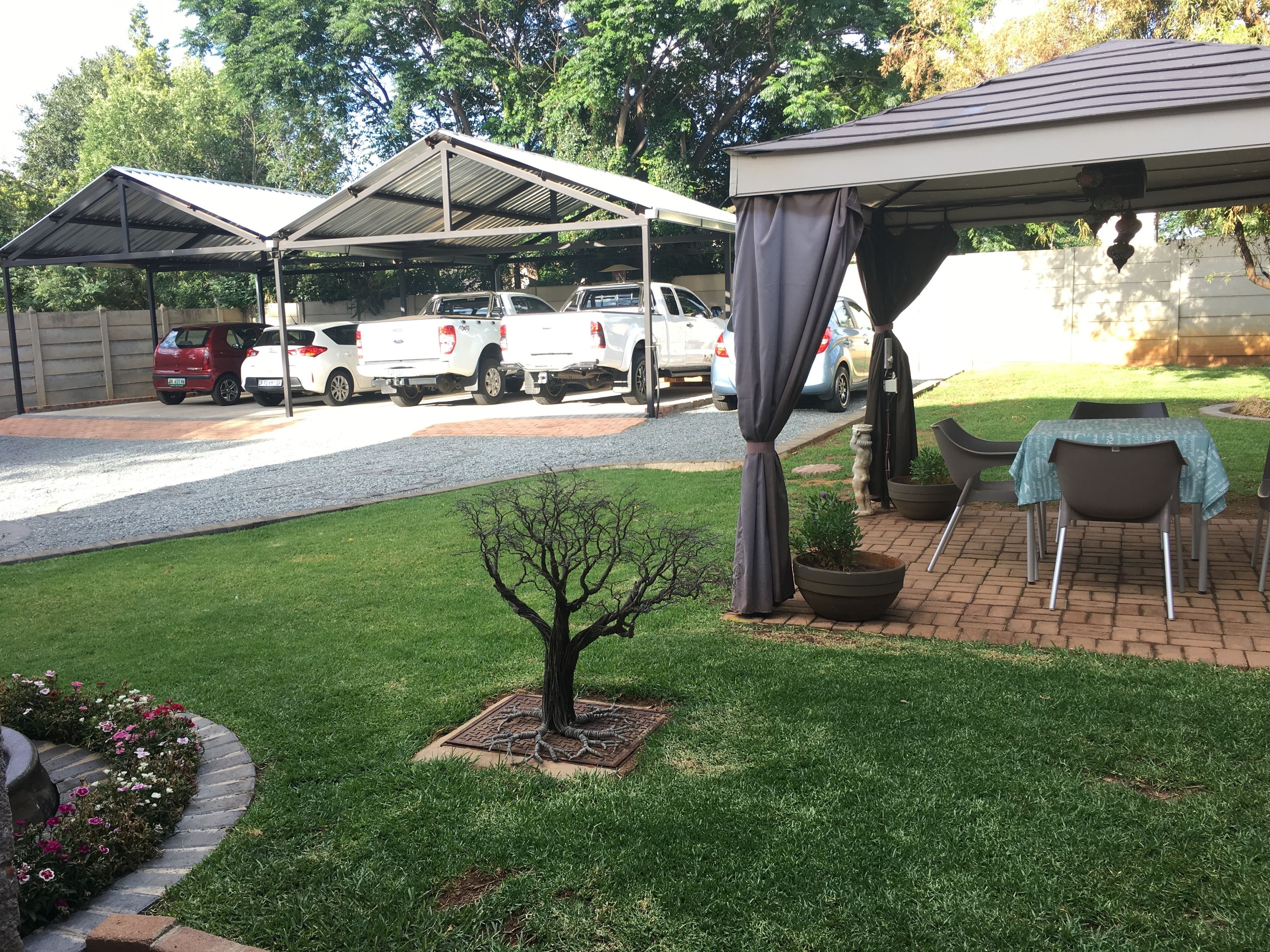 A covered outdoor area with a table and chairs, a small tree, and a parking lot with several vehicles.