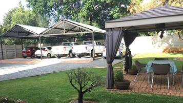 A covered outdoor area with a table and chairs, a small tree, and a parking lot with several vehicles.