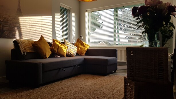 A living room with a dark sofa, yellow cushions, a woven chair, and a large window with blinds.