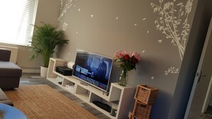 A living room with a flat-screen TV on a white stand, a potted plant, a vase with flowers, and a decorative wall.