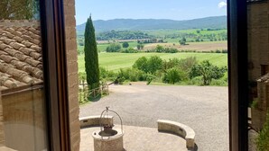 A view from a room with a large window overlooking a countryside landscape, including a stone well and a circular stone structure.