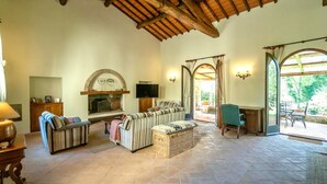 A spacious living room with a fireplace, a sofa set, a television, and a view of the outdoors through large windows.