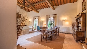 A spacious dining area with a wooden table and chairs, surrounded by windows with curtains, a fireplace, and a bookshelf.