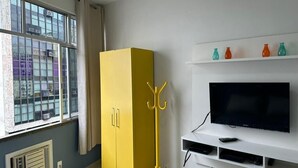 A room with a large window, a yellow wardrobe, a TV on a white stand, and a small shelf with colorful vases. - Rio Habitat - Nsa Sra Apartaments (Rio de Janeiro)