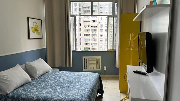A bedroom with a bed, a desk with a computer, a TV, and a window with a view of buildings. - Rio Habitat - Nsa Sra Apartaments (Rio de Janeiro)
