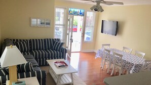 A living room with a striped sofa, a coffee table, a TV mounted on the wall, and a dining area with a table and chairs.