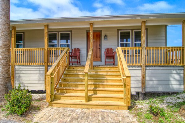 A two-story building with a wooden staircase leading to a porch with two red Adirondack chairs. - Cozy Pet Friendly Duplex Cottage nestled right in the heart of Port Aransas! (Port Aransas)