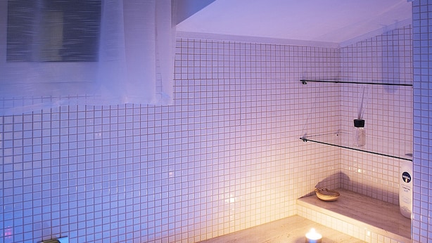 Een badkamer met een bad, betegelde wanden, een raam met gordijnen en een plank met toiletartikelen.