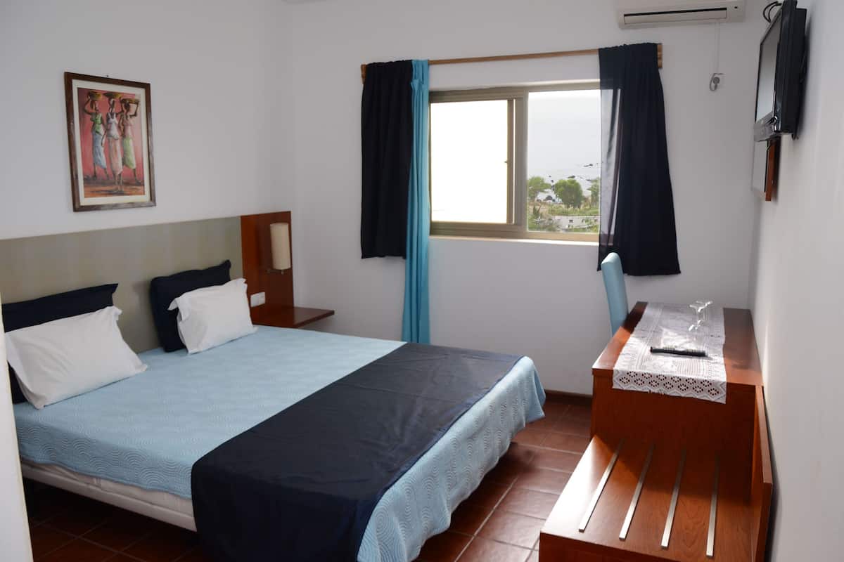 A bedroom with a bed, a desk with a chair, a window with curtains, a painting on the wall, and an air conditioning unit.