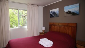 Quarto de hotel com uma cama grande, cabeceira de madeira, janela com cortinas, mesa de cabeceira com abajur e duas fotos emolduradas na parede.
