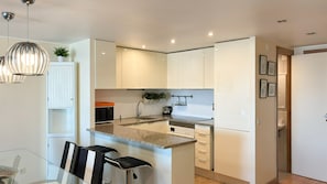 A modern kitchen with white cabinetry, a granite countertop, and a dining area with a glass table and chairs. - Vista Marina Exclusiva | T1 HsRentals Vilamarina (Vilamoura)