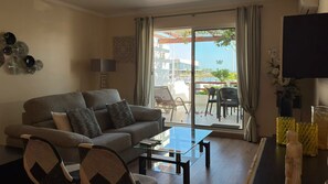 Ein Wohnzimmer mit Sofa, Sesseln, einem Couchtisch, einem Fernseher und Blick auf eine Marina durch große Fenster.