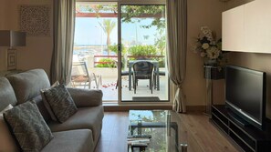A living room with a sofa, a glass coffee table, a flat-screen TV, and a view of a balcony with outdoor furniture.