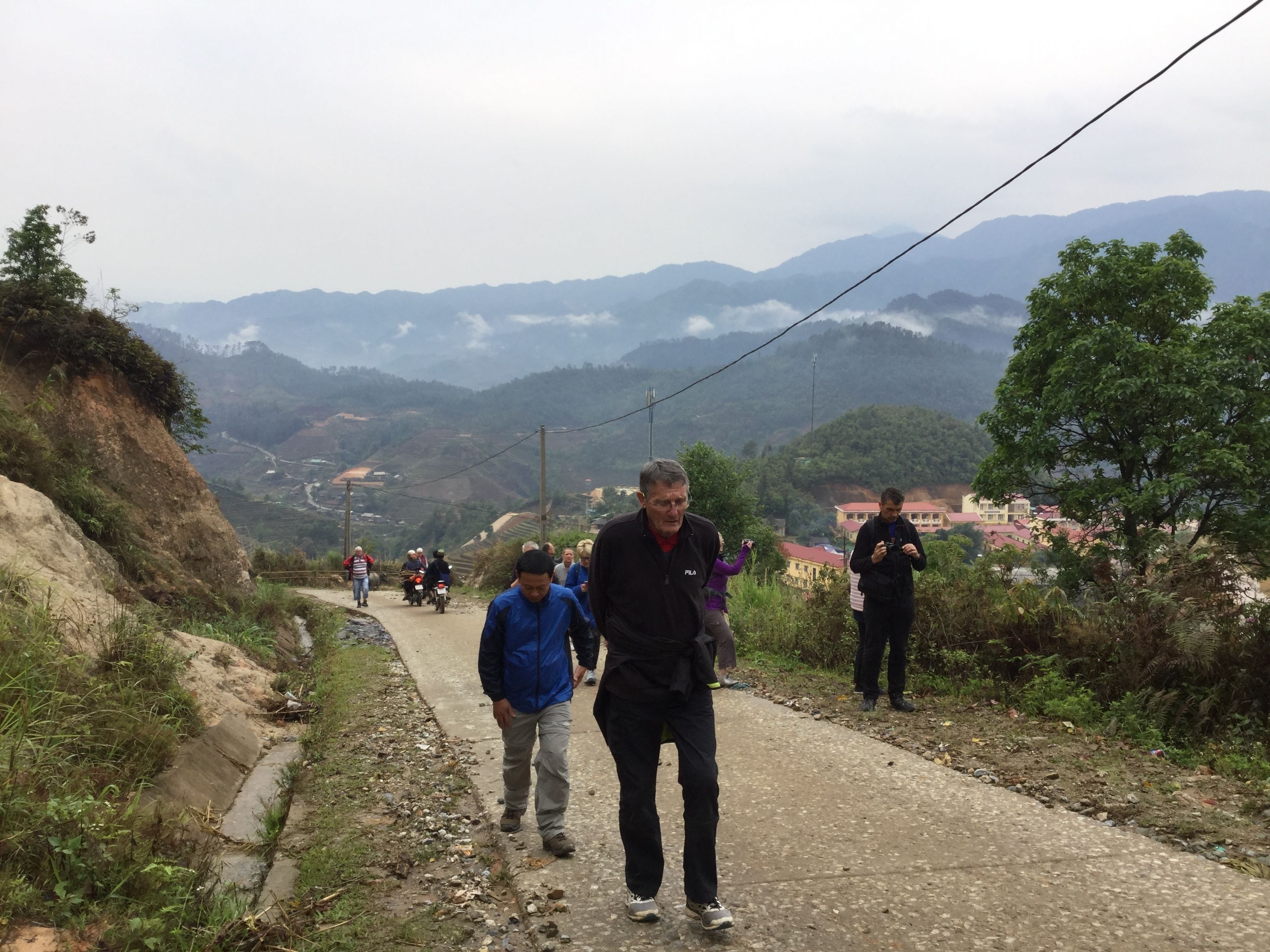 A paved road with people walking and riding motorcycles, surrounded by greenery and distant mountains.