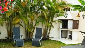 A backyard with two lounge chairs, a small barbecue area, and lush tropical plants.