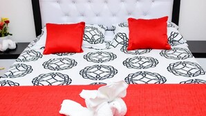 A neatly made bed with a red bedspread and two red pillows, a white tufted headboard, and a small bedside table with a vase of flowers.