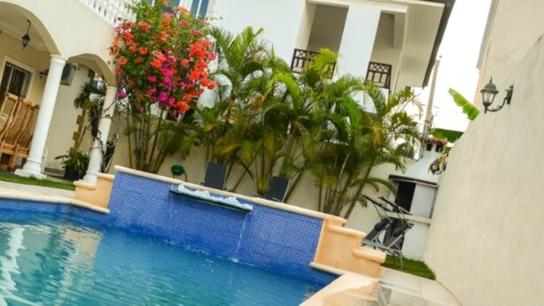 A swimming pool with a tiled edge, a patio area with wicker furniture, and a building with a white facade and columns.