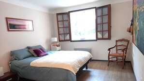 A bedroom with a bed, a chair, a window with open shutters, a bedside table with a lamp, and a painting on the wall.