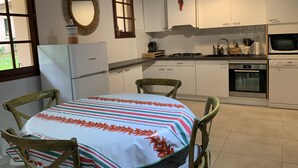 A kitchen with white cabinets, a stove, an oven, and a refrigerator. There is a dining table with chairs and a window with a view of the outdoors.