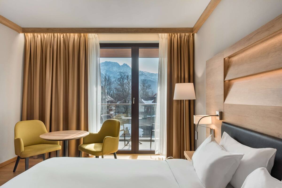 Habitación de hotel con una cama, dos sillas, una mesita, una lámpara y vistas a las montañas a través de un ventanal grande.
