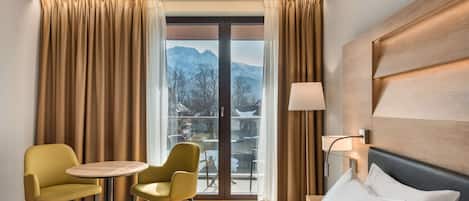 A hotel room with a bed, two chairs, a small table, a lamp, and a view of mountains through a large window.
