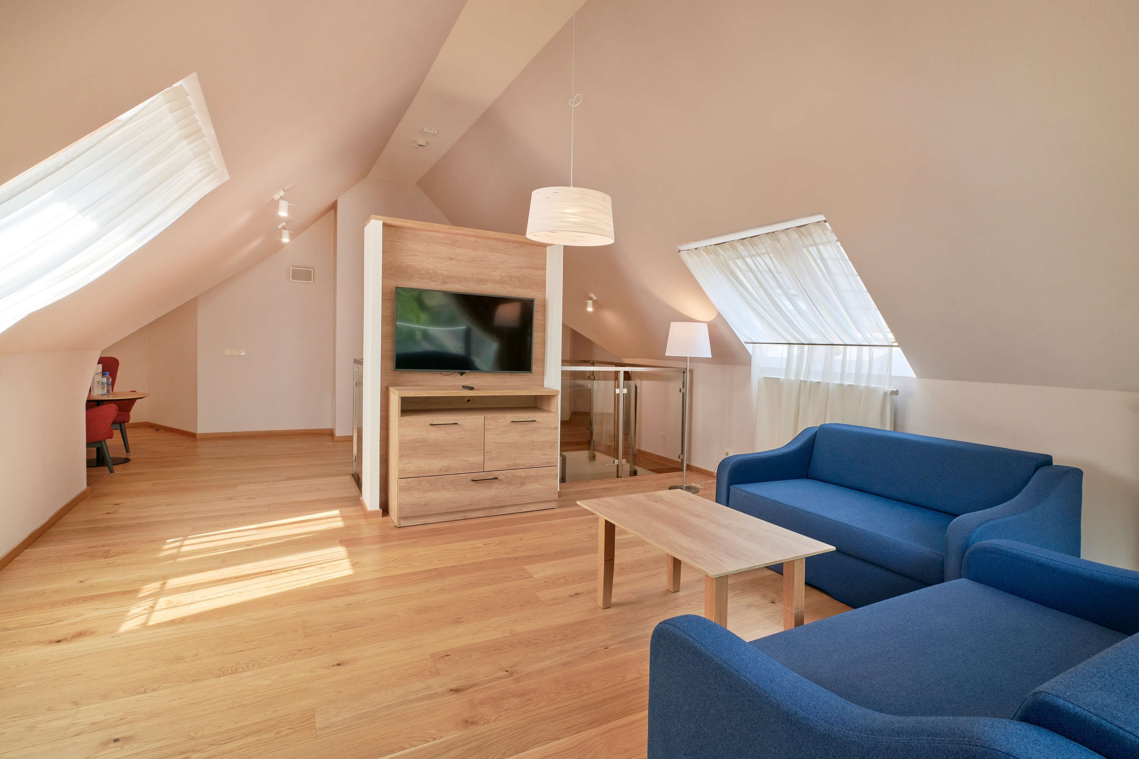 A modern loft apartment with a sloped ceiling, wooden flooring, a blue sofa, a wooden cabinet, a TV, and large windows with sheer curtains.