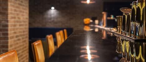 Zona de bar con una barra larga, múltiples lámparas colgantes y una pared de ladrillo.
