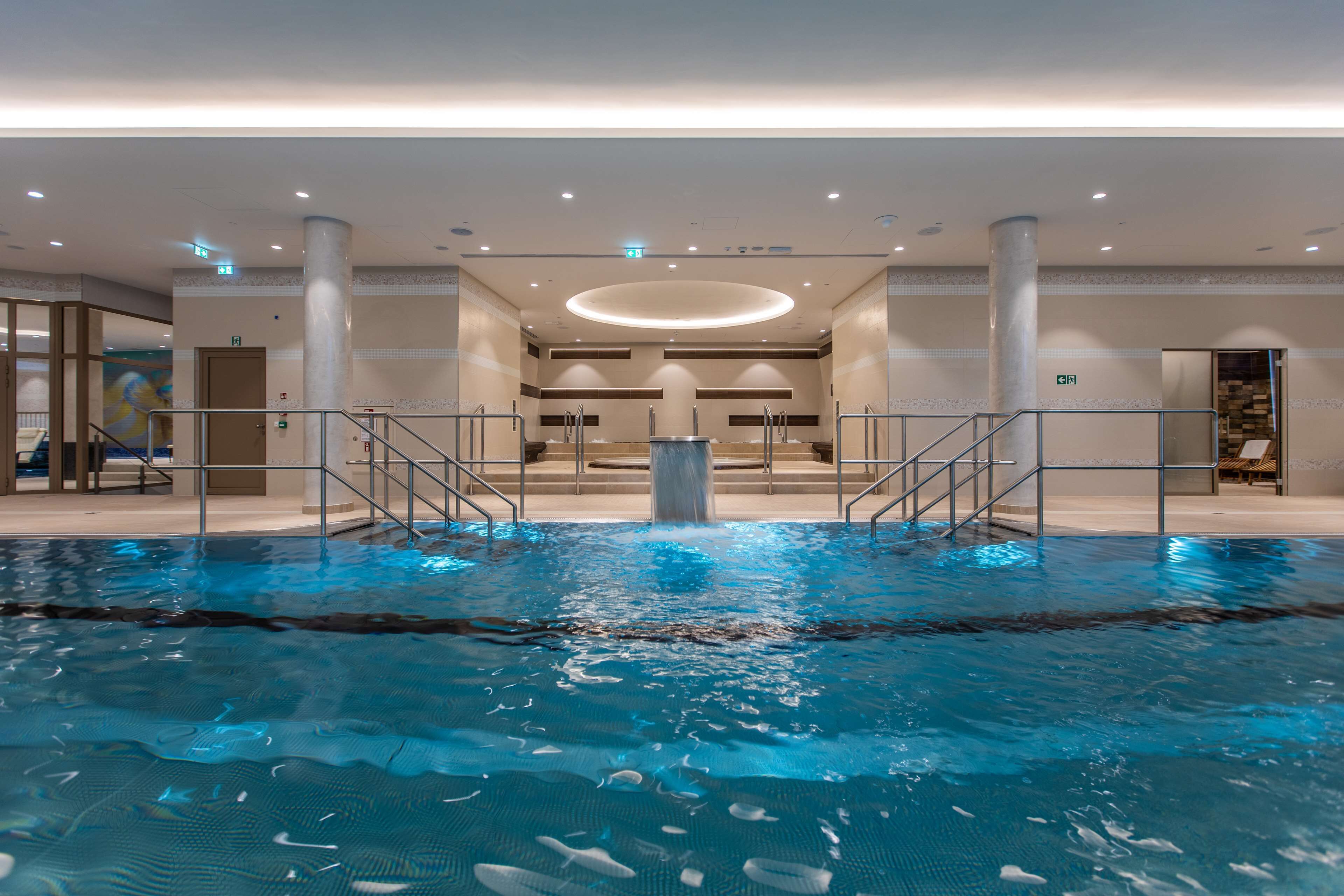 Piscina interior con diseño moderno, rodeada por un amplio vestíbulo con techo alto y una lámpara circular central.