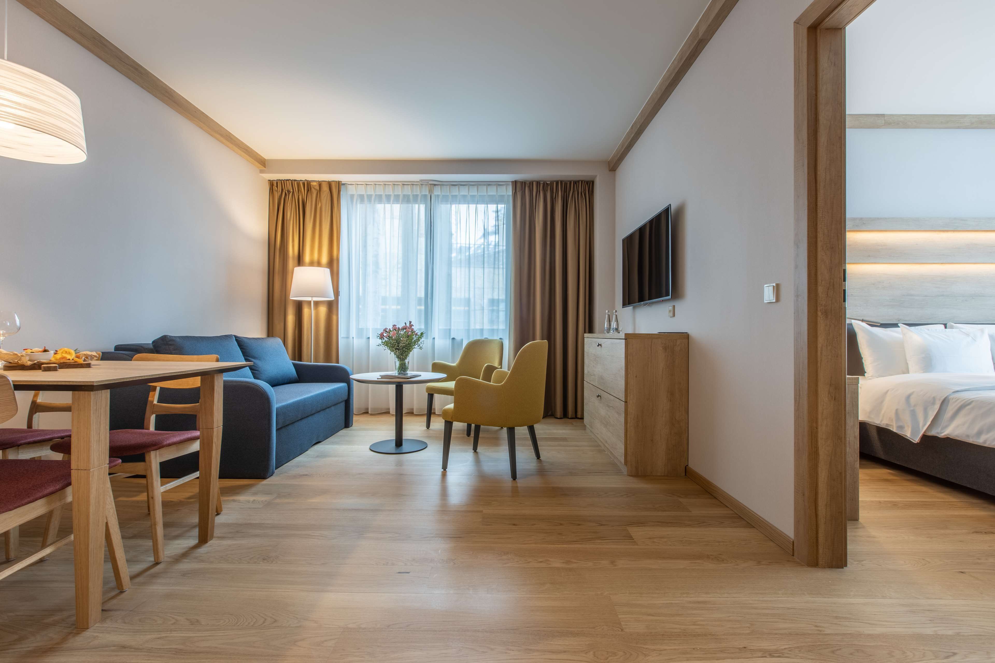 Habitación de hotel con piso de madera, sofá, mesa de comedor con sillas, una mesita redonda con lámpara, televisor montado en la pared y cama con cabecera.