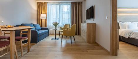 Habitación de hotel con piso de madera, sofá, mesa de comedor con sillas, una mesita redonda con lámpara, televisor montado en la pared y cama con cabecera.