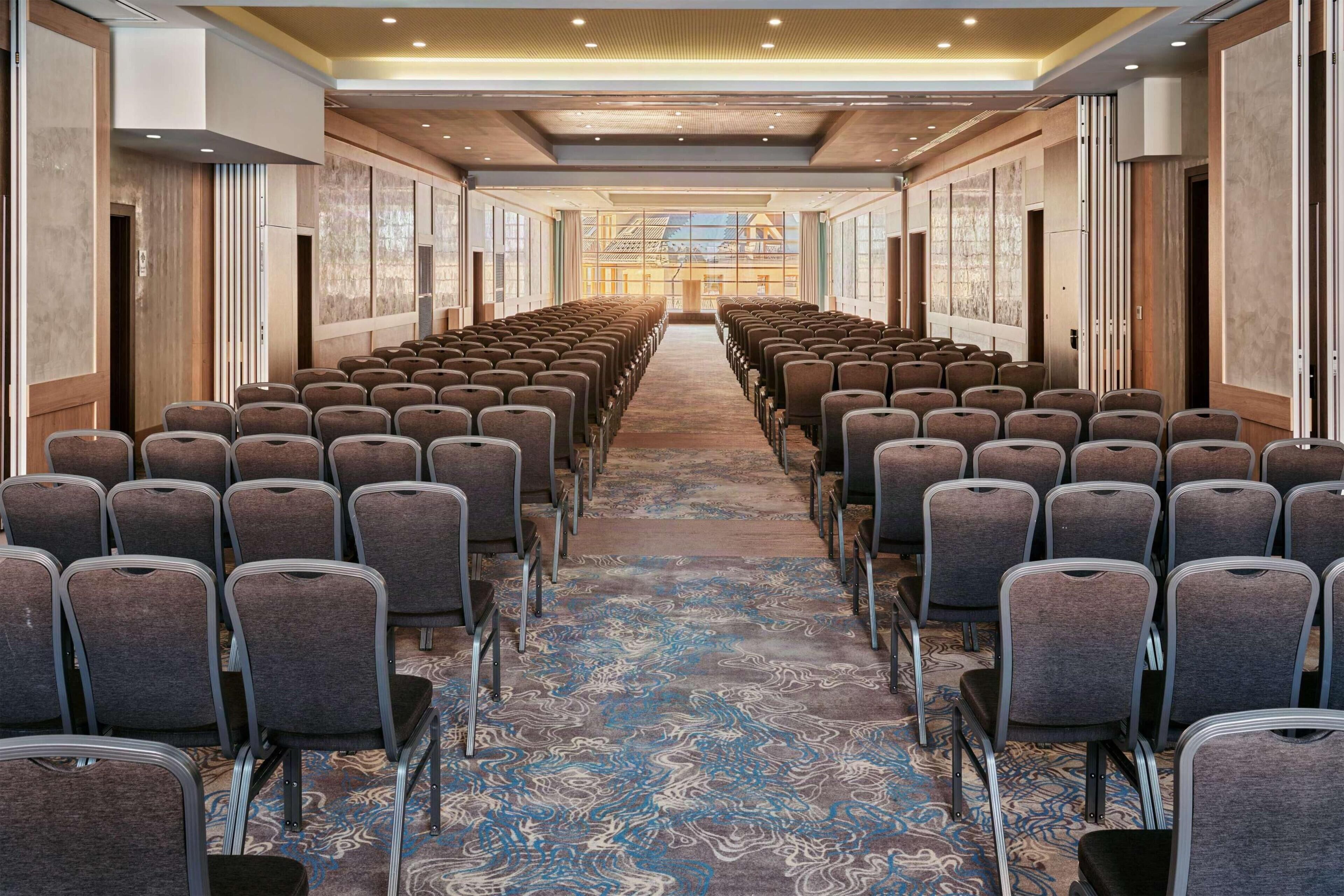 Un salón de conferencias con filas de sillas, una alfombra estampada y grandes ventanas con vistas a un edificio.