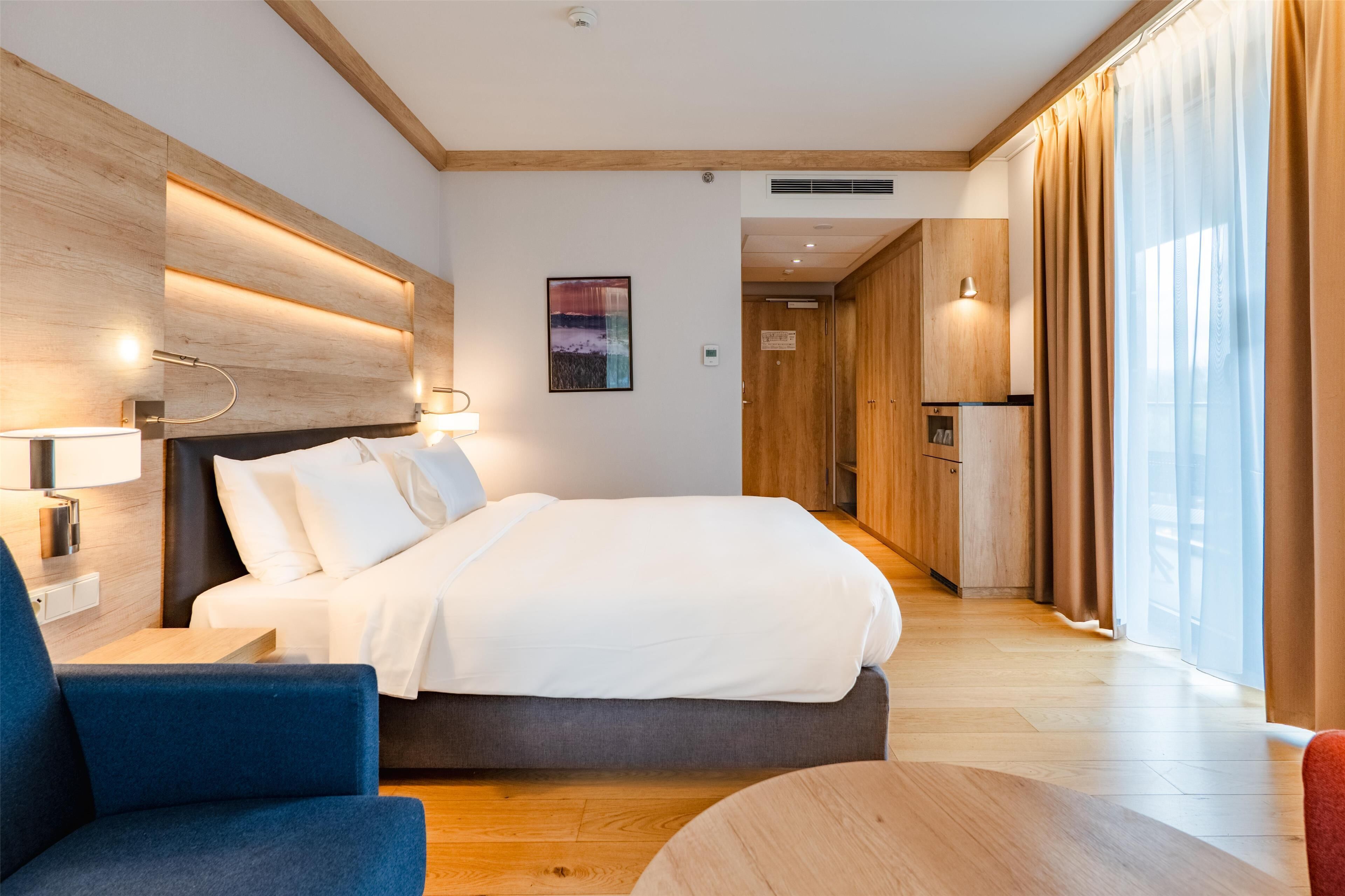 A modern hotel room with a large bed, a desk, and a chair. There is a wooden headboard, a wall-mounted lamp, and a painting on the wall.