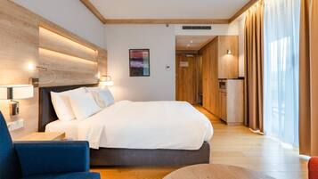 A modern hotel room with a large bed, a desk, and a chair. There is a wooden headboard, a wall-mounted lamp, and a painting on the wall.