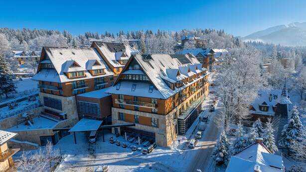 A large, multi-story hotel with a modern architectural design, featuring a mix of brick and glass facades, and a parking area with several cars. - Radisson Blu Hotel & Residences, Zakopane (Zakopane)