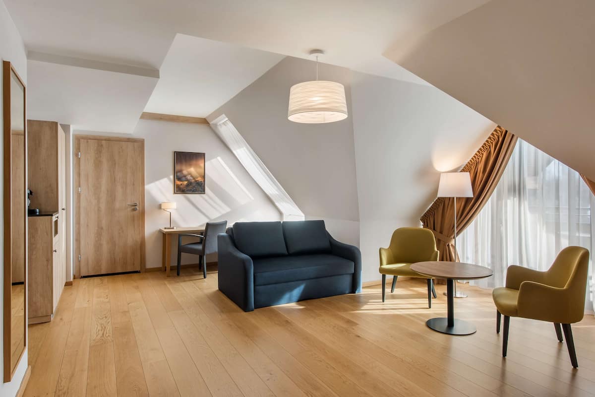 Una habitación moderna estilo loft, con piso de madera, sofá azul oscuro, sillas amarillas, una pequeña mesa redonda y un gran ventanal con cortinas.