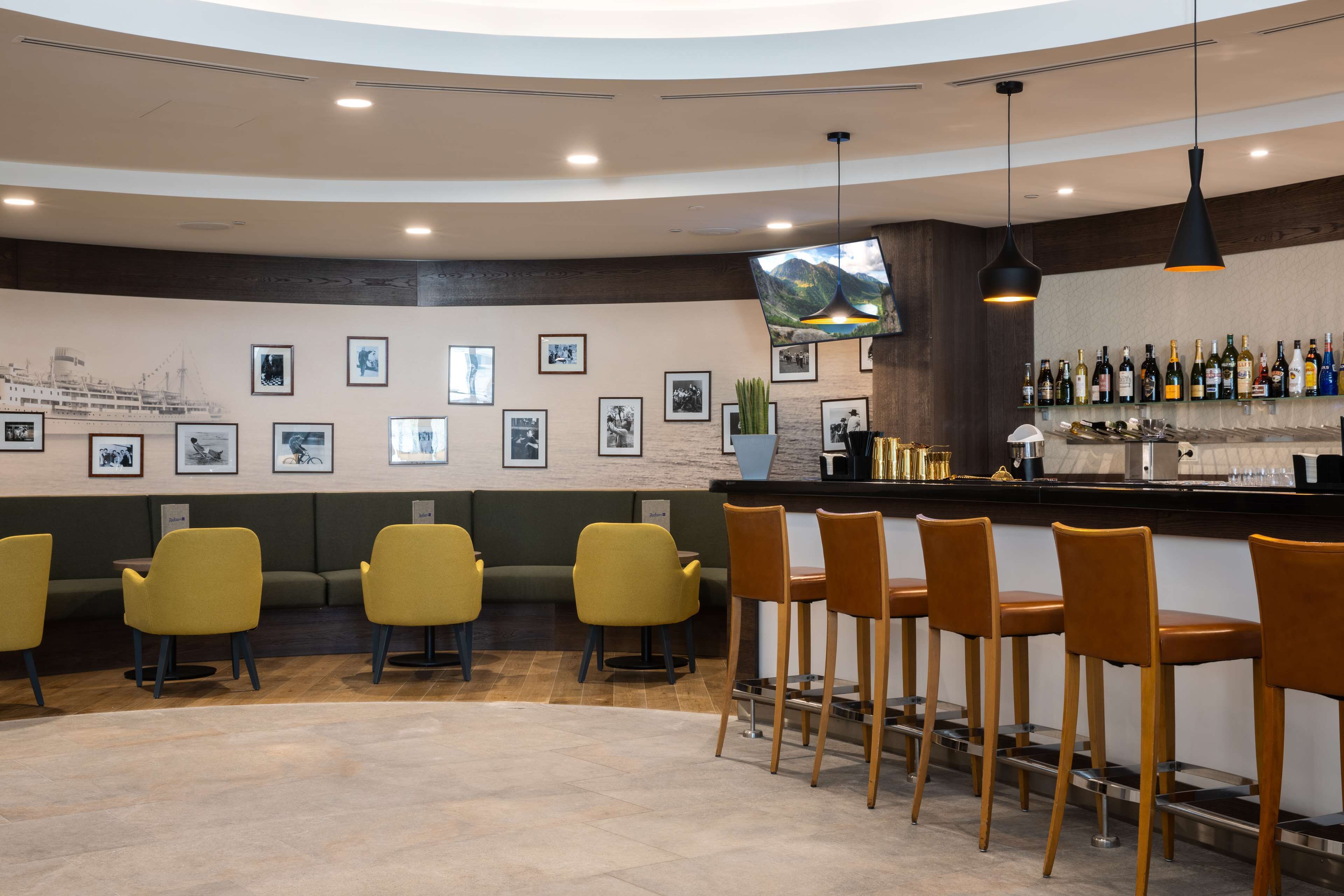 Een bar met gele stoelen, een toonbank met flessen en ingelijste foto's aan de muur.