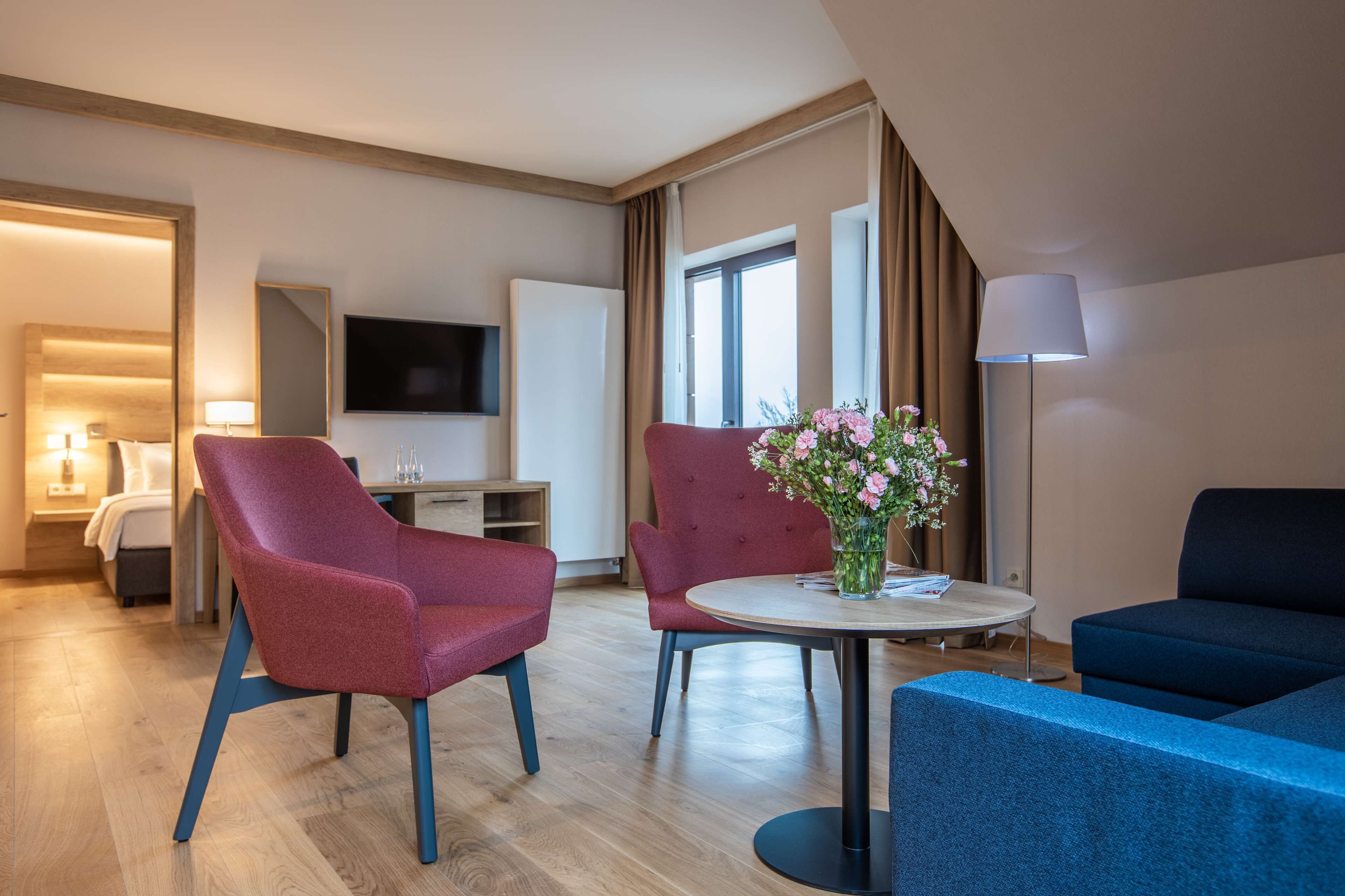 A modern hotel room with a wooden floor, a round dining table with chairs, a sofa, a TV, and a lamp.